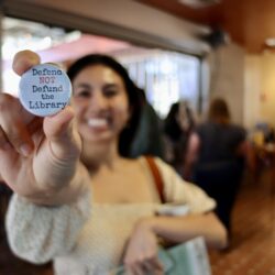 Image contains: A San Diego Book Crawler holds up a 'Defend NOT Defund the Library' pin.