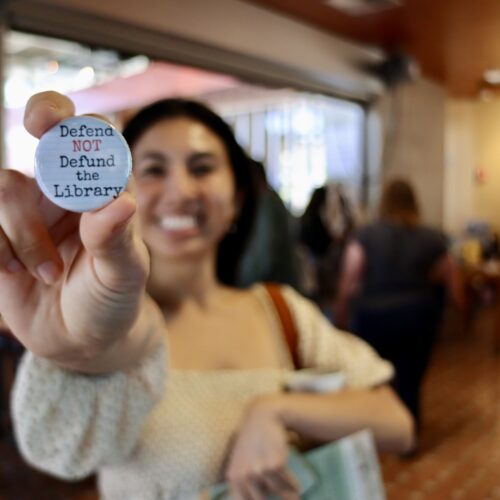 Image contains: A San Diego Book Crawler holds up a 'Defend NOT Defund the Library' pin.