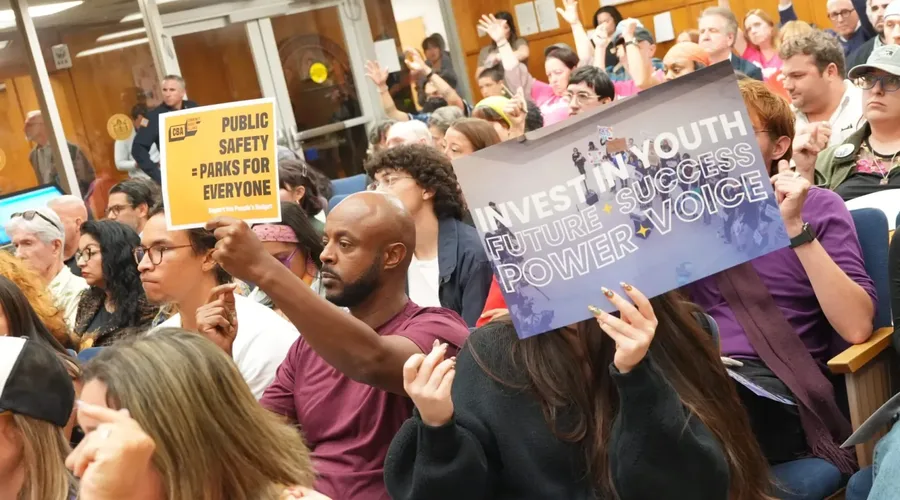 Petros Hagos, left, and Claire Snyder, right, hold posters during a budget hearing at City Hall on Wednesday, May 27, 2026, in San Diego. (Michael Ho / For The San Diego Union-Tribune)